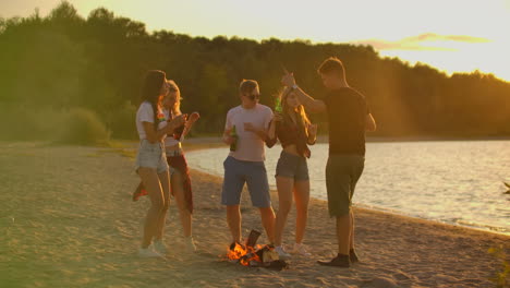 The-students-are-dancing-in-shorts-and-t-shirts-around-bonfire-on-the-sand-beach-with-beer.-They-are-enjoying-the-summer-evening-on-the-river-coast-on-the-open-air-party.